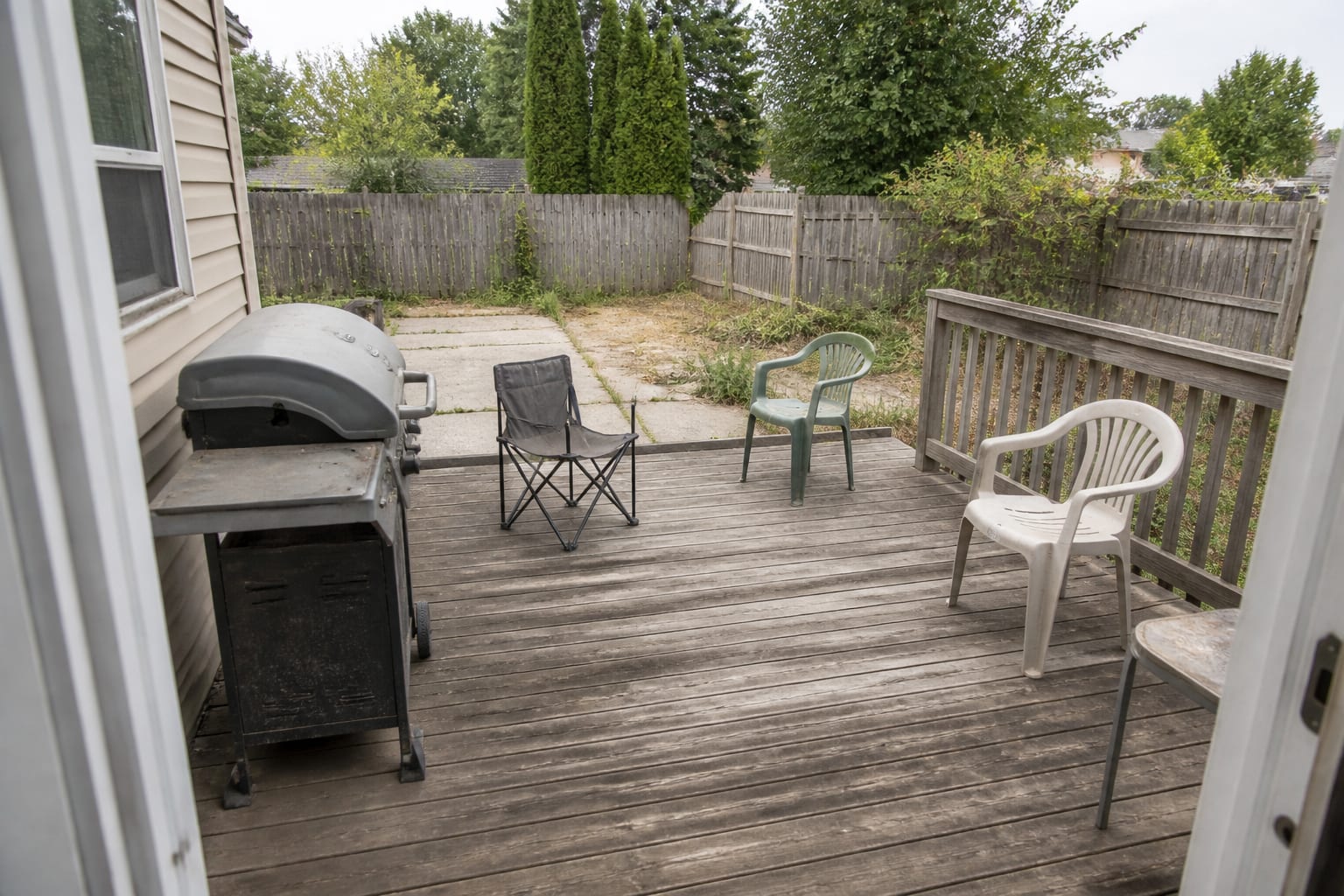 Weathered backyard deck with mismatched chairs, bare railings, crowded grill placement, no shade, and undefined seating areas.