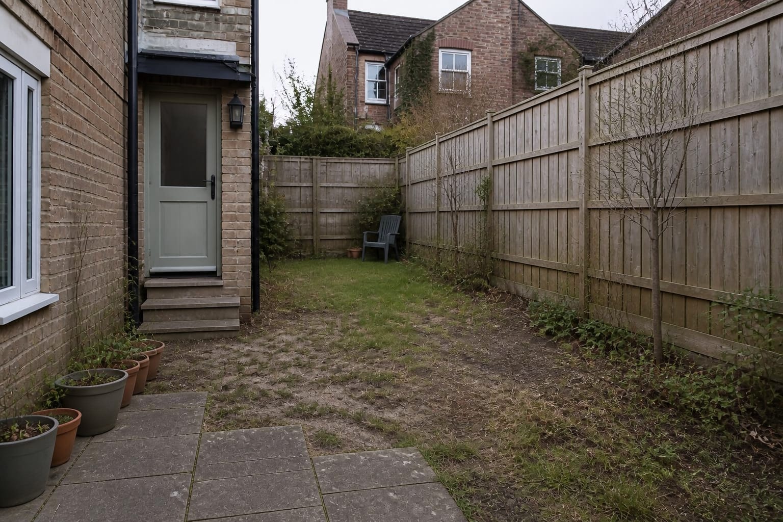 neglected garden with patchy lawn, shallow border, scattered small pots, bare fence, and no clear path or seating destination.