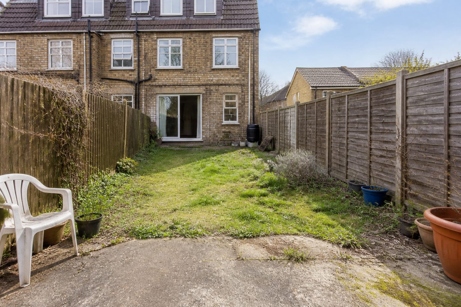 neglected garden with patchy lawn, shallow border, scattered small pots, bare fence, and no clear path or seating destination.
