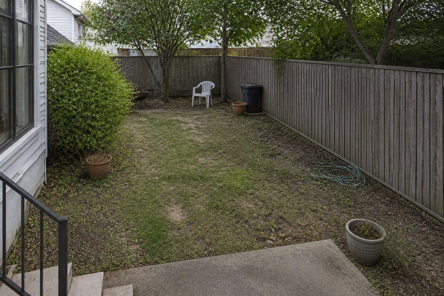 Patchy residential garden with thin lawn, undefined planting edges, bare fence, scattered pots, and no clear route through the outdoor space.