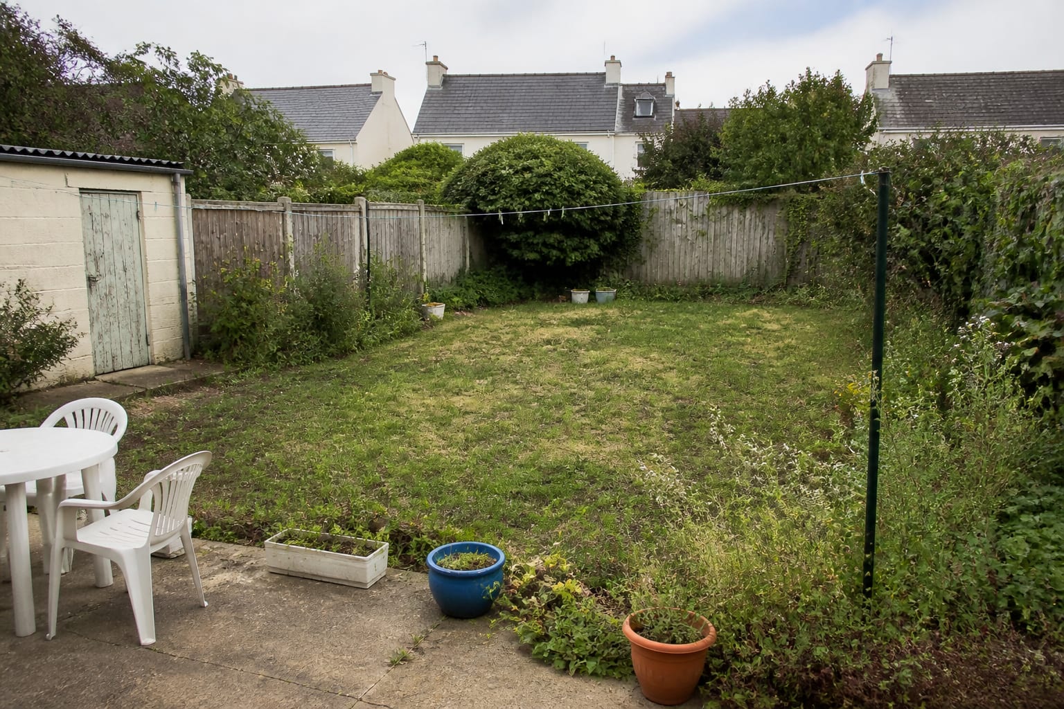 Patchy residential garden with thin lawn, undefined planting edges, bare fence, scattered pots, and no clear route through the outdoor space.