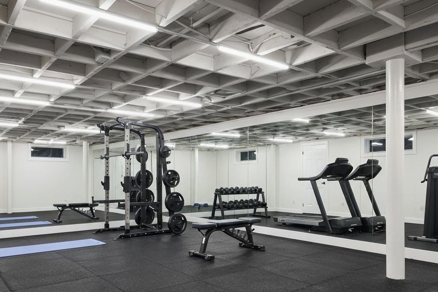 finished basement home gym with rubber flooring, warm lighting, wall mirrors, compact equipment zones, and closed storage