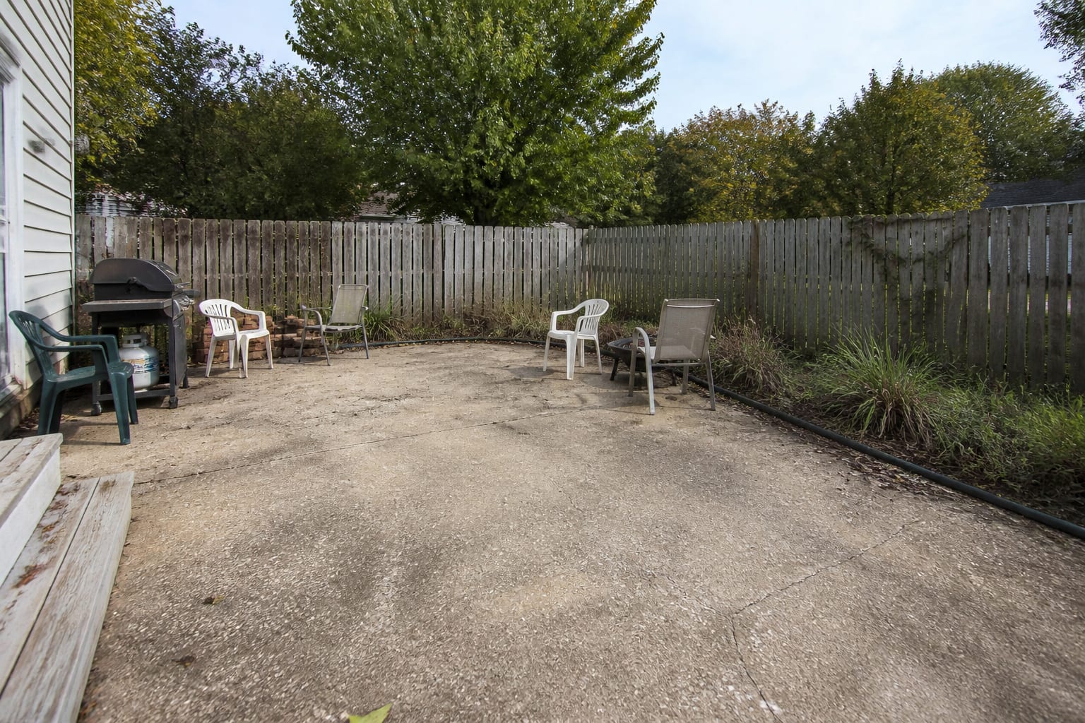 under designed patio with exposed concrete, no shade, scattered chairs, visible fence line, and a back door path interrupted by loose furniture.