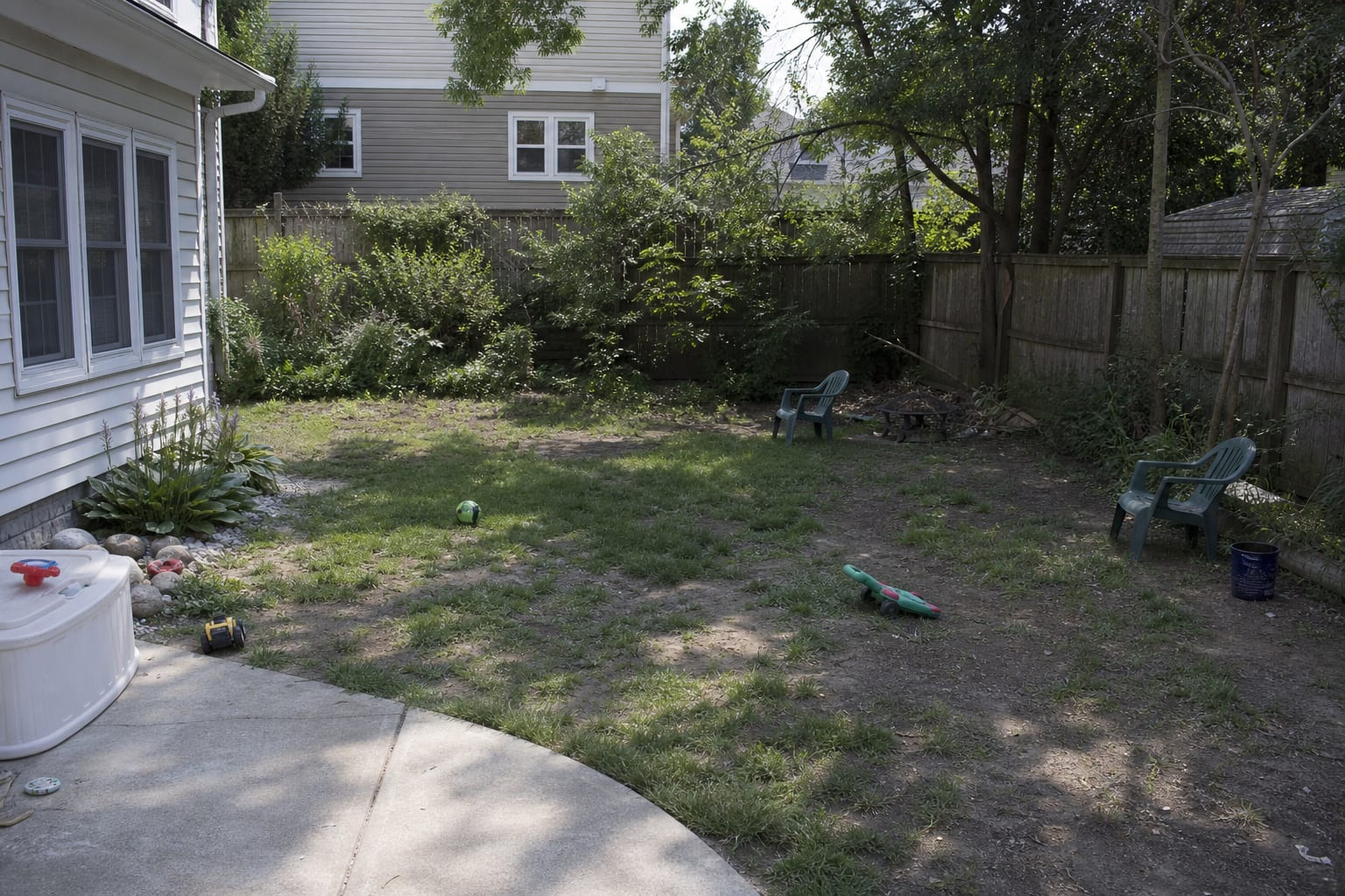 Patchy dry backyard lawn with bare soil, uneven shade, scattered toys, and no clear edge between grass and planting beds.