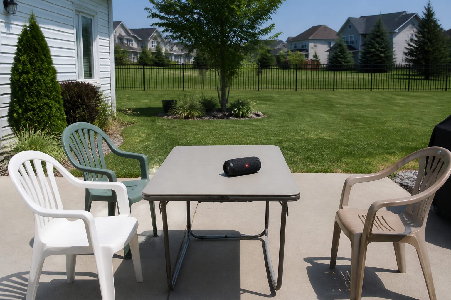 Plain open backyard patio with a portable Bluetooth speaker, mismatched patio chairs, and no shade structure or TV