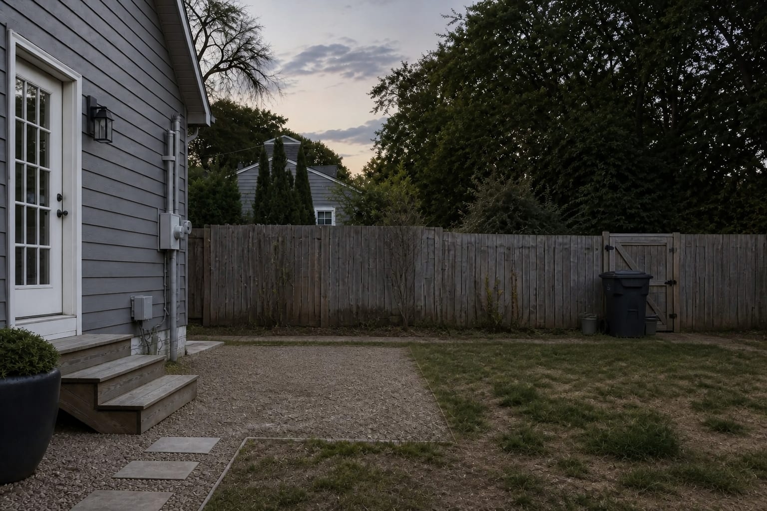 backyard with an uneven faded fence, exposed trash area, patchy grass, and no planting to soften the property line