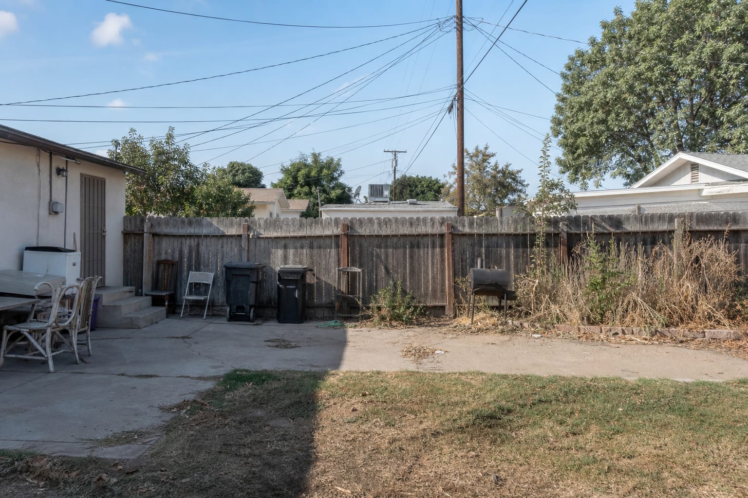 backyard with an uneven faded fence, exposed trash area, patchy grass, and no planting to soften the property line