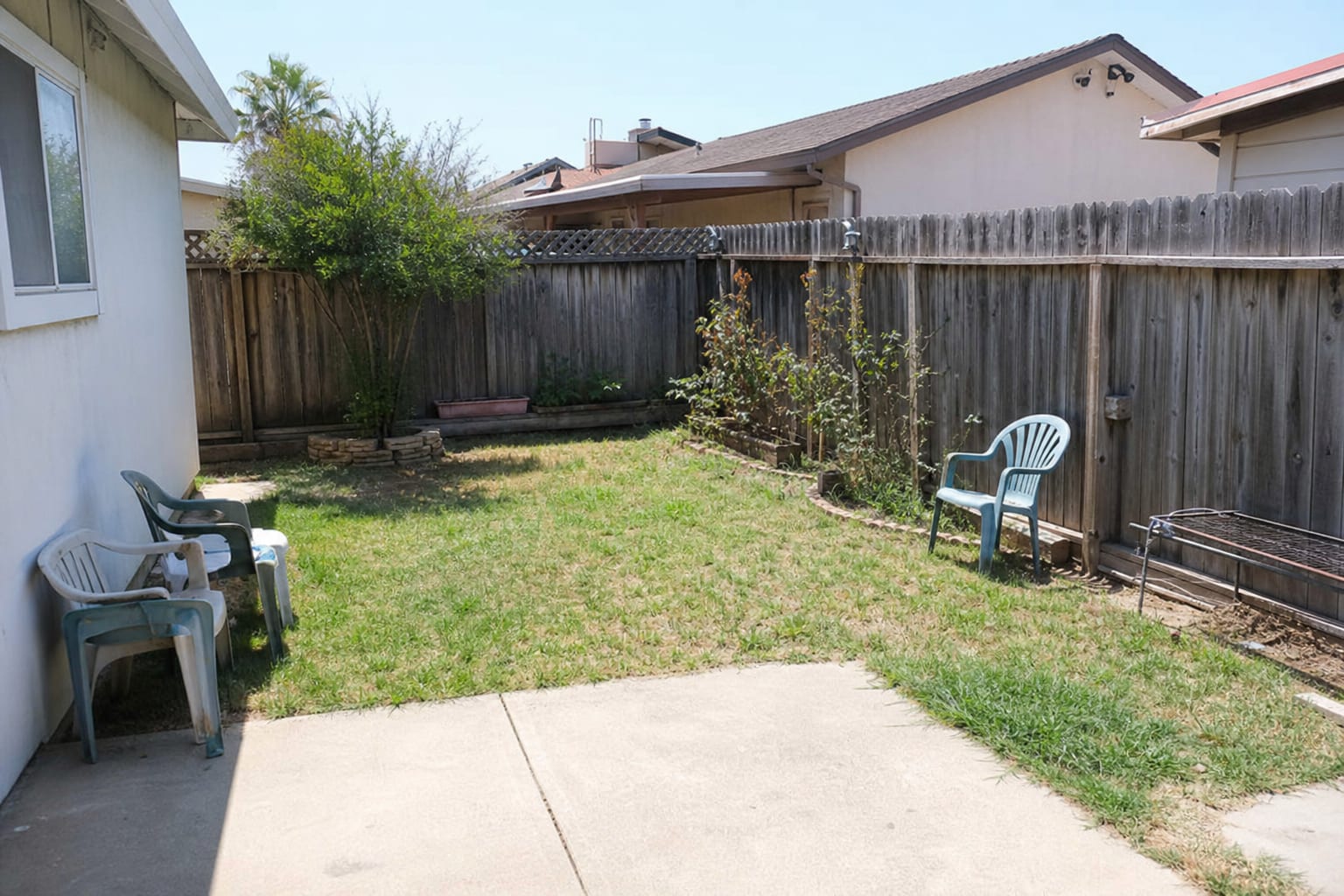 Plain backyard with patchy grass, mismatched plastic chairs, exposed fence lines, and no defined place to sit.