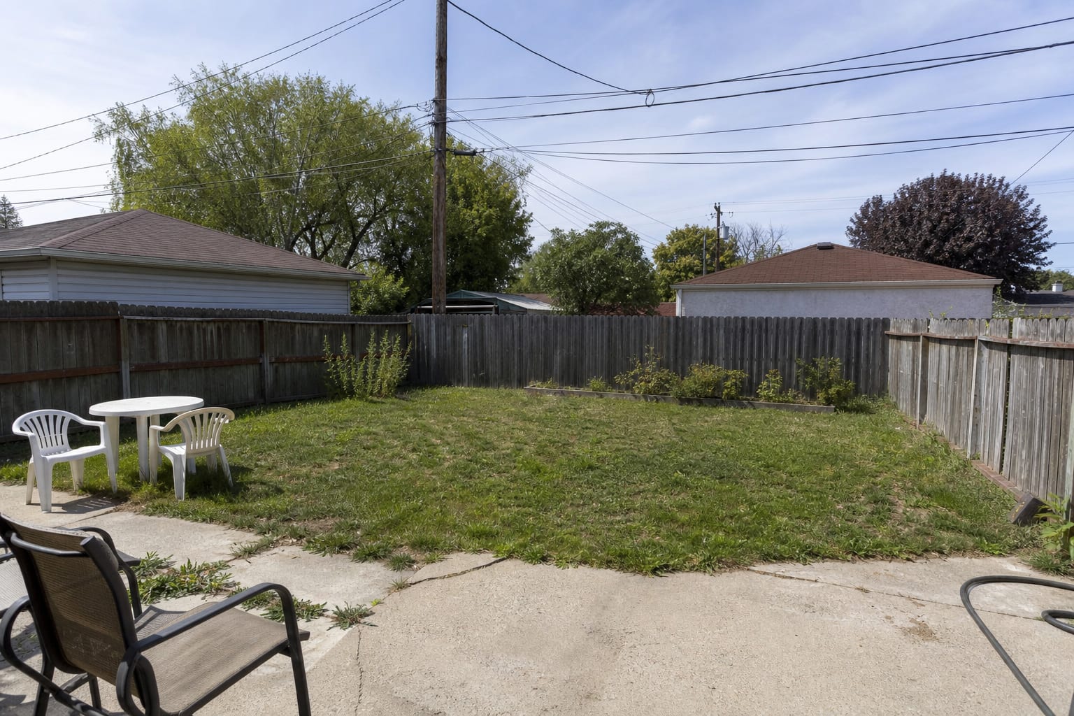 A flat backyard lawn with no shade, no pool layout, scattered chairs, and a fence line that feels exposed and unfinished.