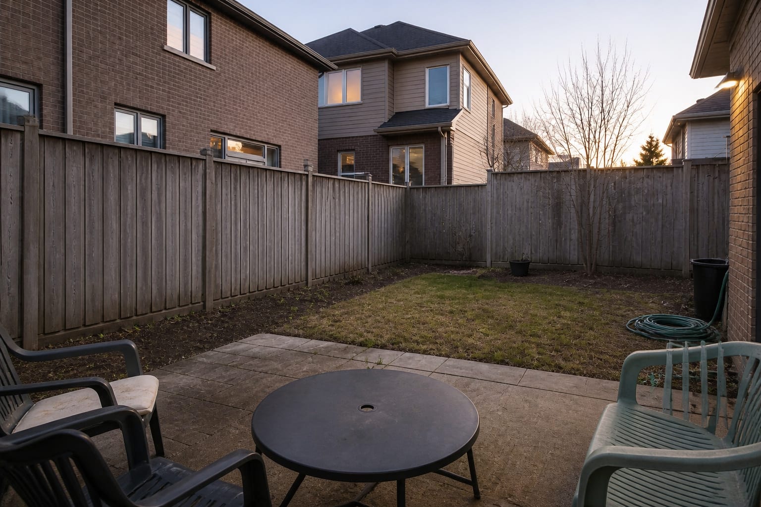 Exposed backyard seating area with a plain fence, sparse lawn, visible neighboring windows, and no layered planting.