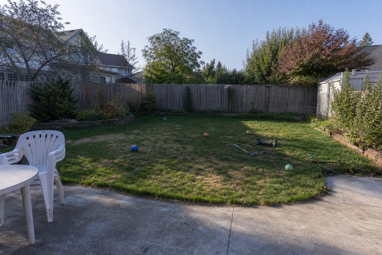 Under-designed backyard lawn with patchy grass, no active play zone, exposed fence line, and scattered outdoor toys near the patio.