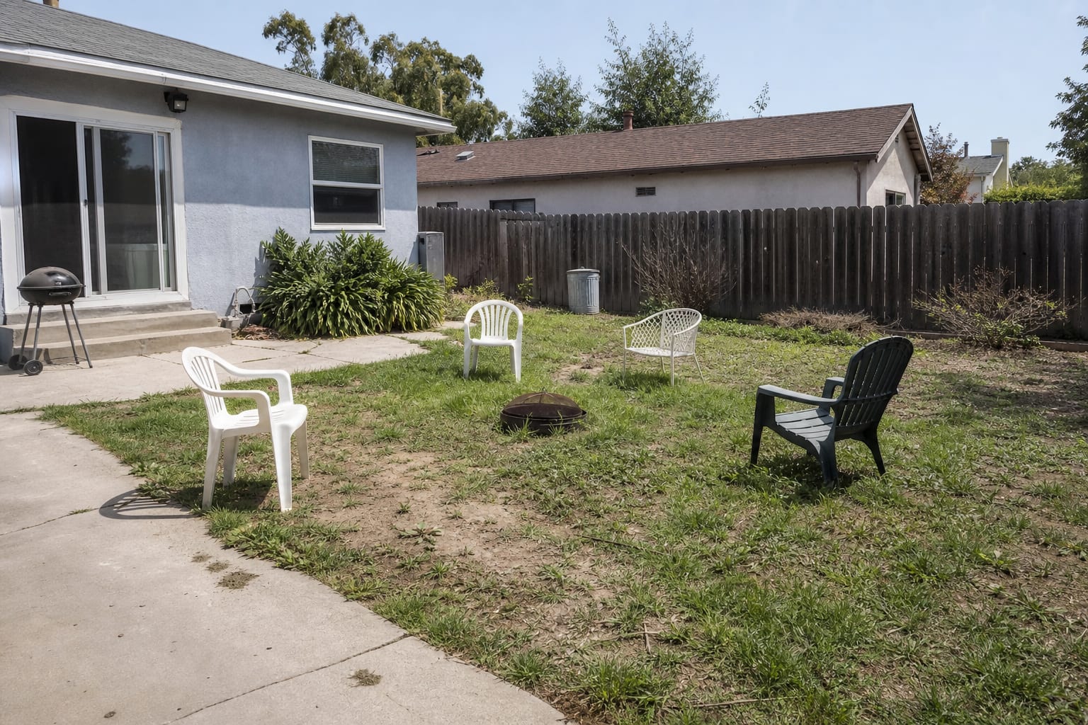 undefined backyard with scattered chairs on patchy lawn, no clear path, and an empty fence line behind the house