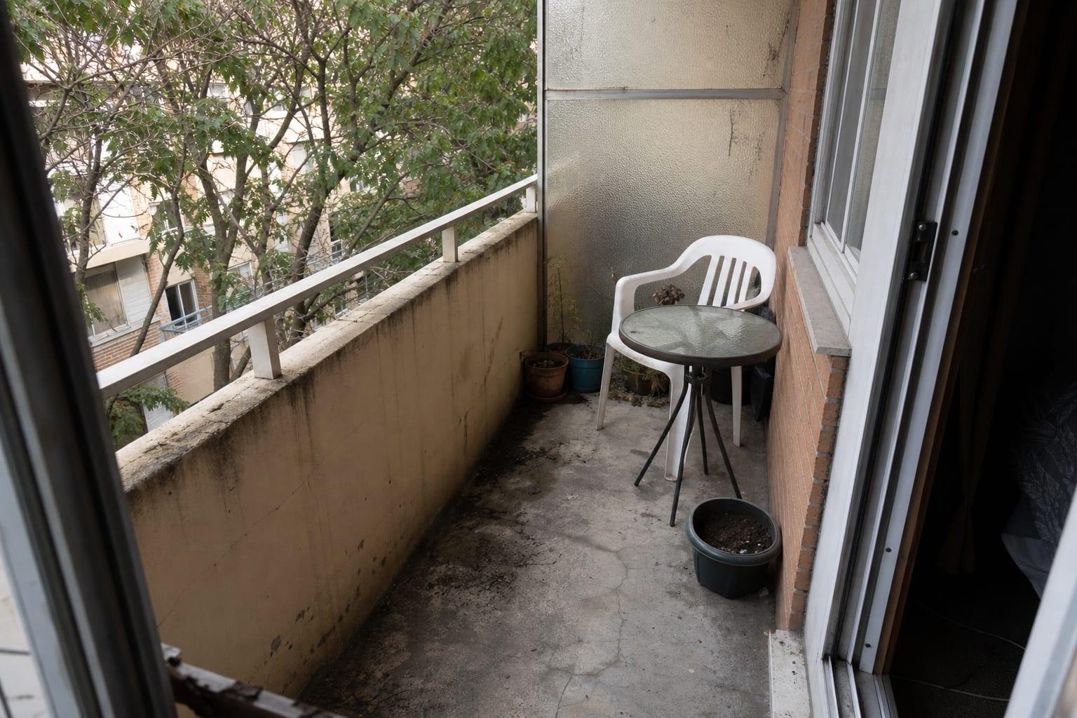 plain concrete apartment balcony with a stained slab, exposed railing posts, and mismatched small outdoor pieces