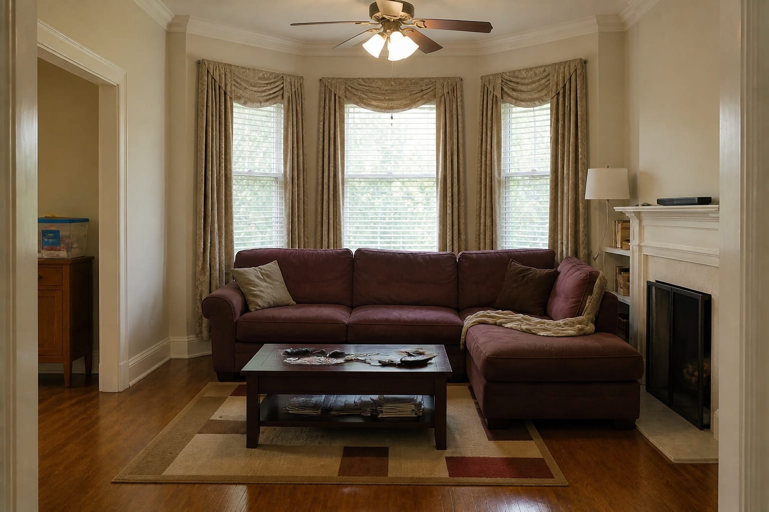 Living room with a maroon microfiber sectional shoved against a three-window bay, heavy formal beige drapes, and a cold ceiling-fan light.