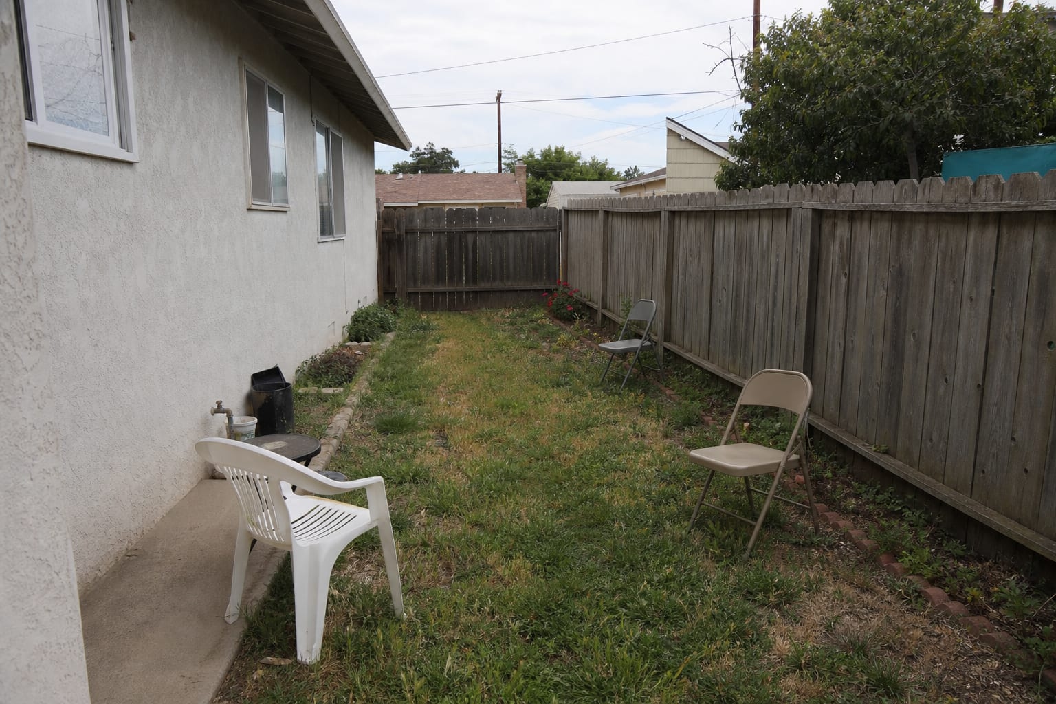 narrow unused backyard side lawn with patchy grass, loose chairs, and no clear recreation zone beside a fence