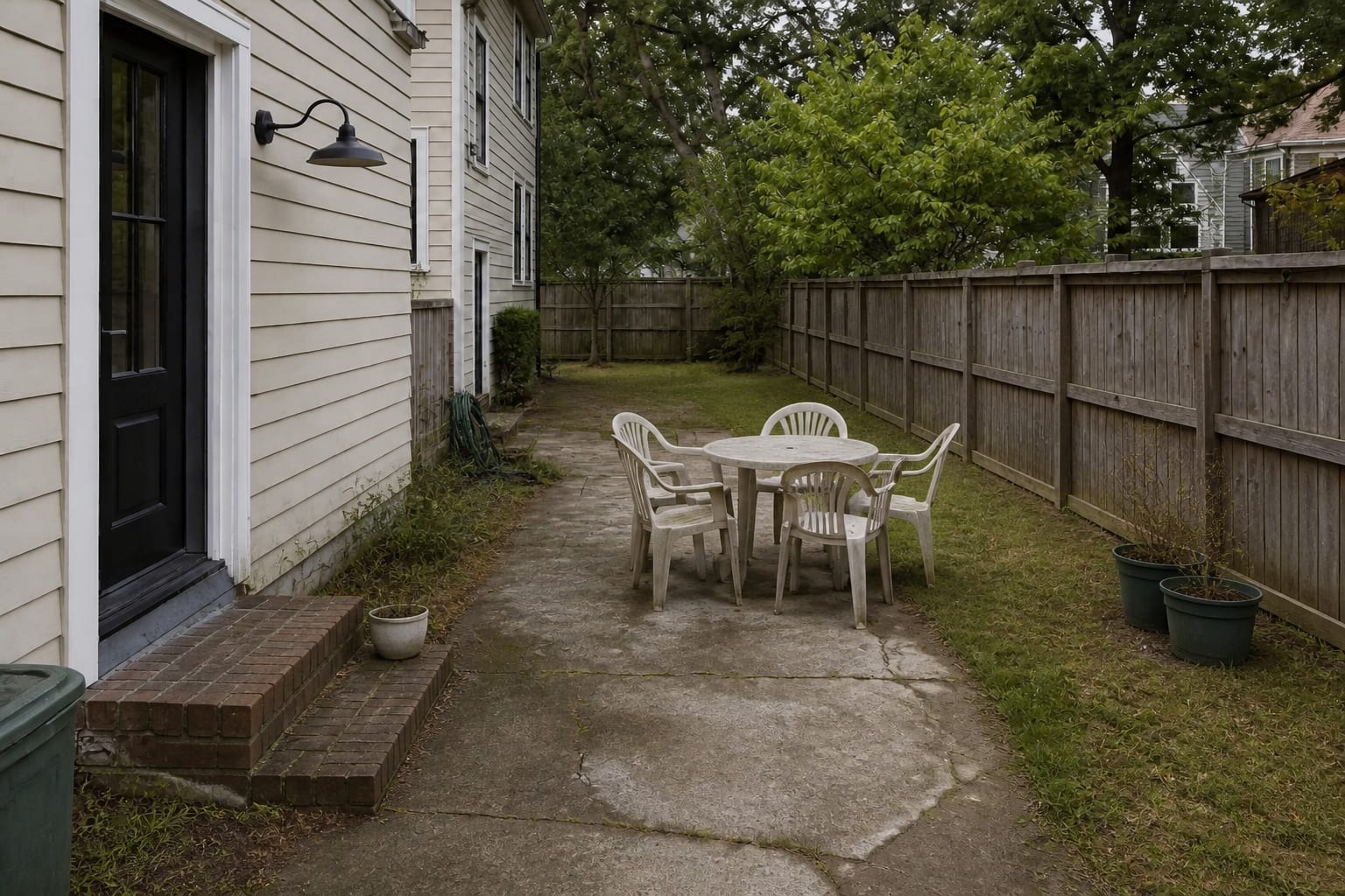 Uneven concrete and thin grass beside an older house with scattered chairs, no patio border, and a blank fence line.