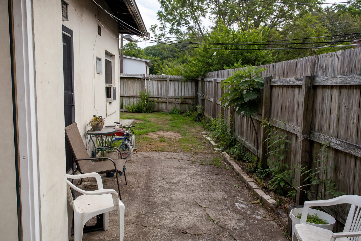 Uneven concrete and thin grass beside an older house with scattered chairs, no patio border, and a blank fence line.