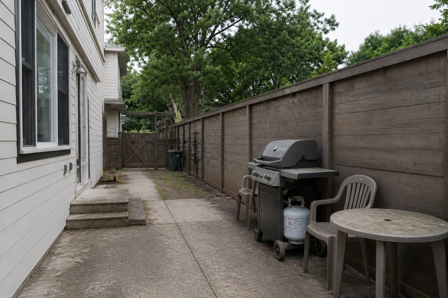 freestanding grill on a plain patio with no counter space, exposed propane tank, scattered chairs, and a narrow path to the house