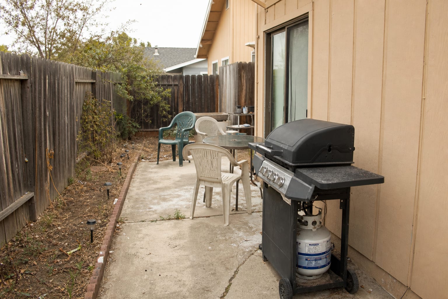freestanding grill on a plain patio with no counter space, exposed propane tank, scattered chairs, and a narrow path to the house