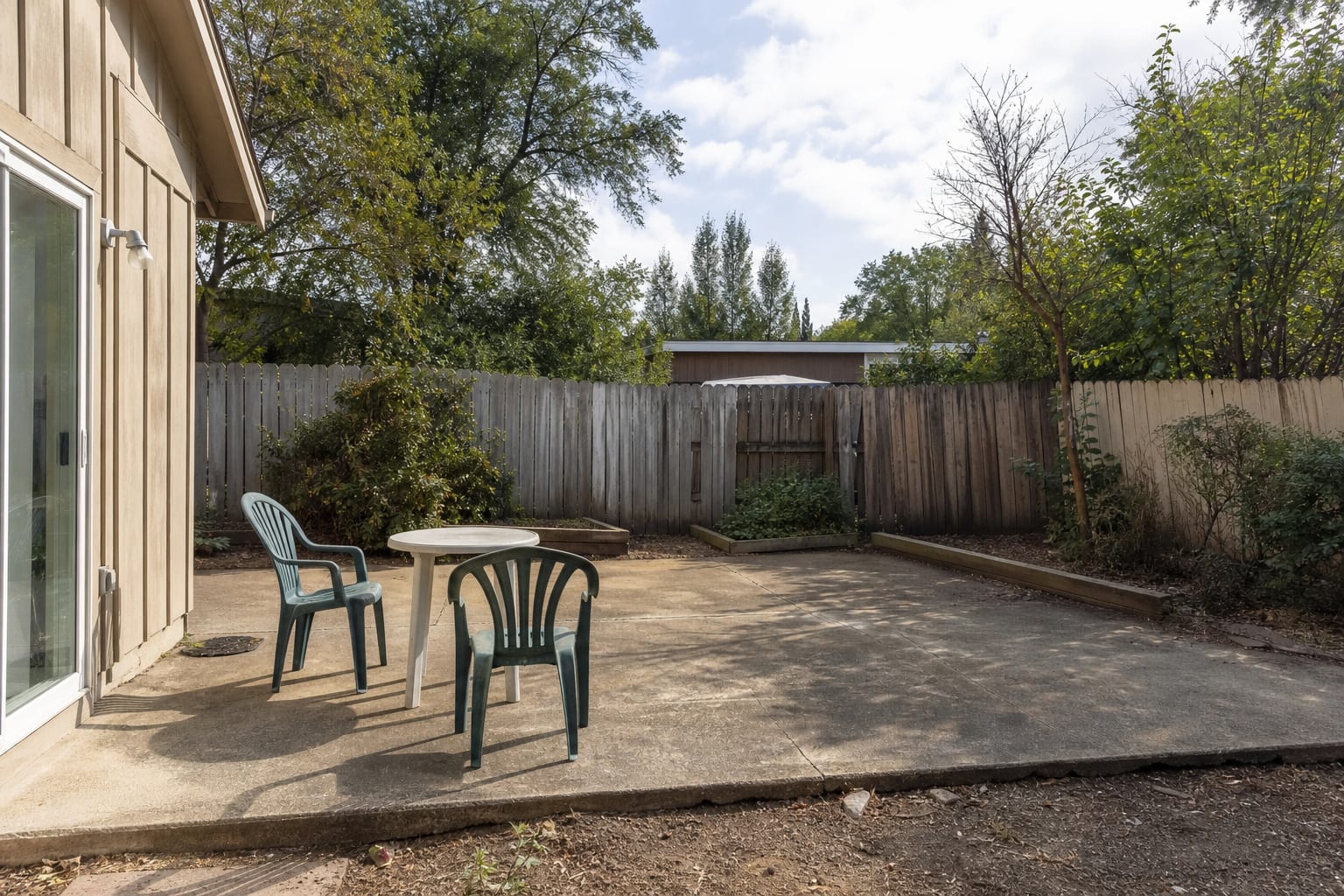 plain gray backyard concrete patio with mismatched chairs, exposed lawn edges, no shade, and a blank house wall.