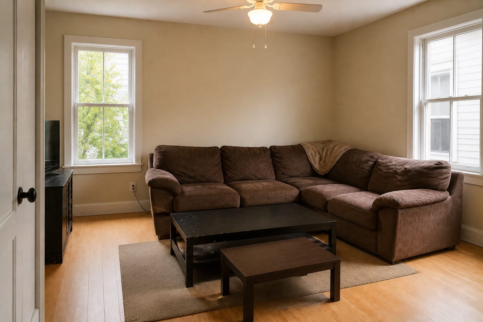 Square living room with an oversized brown microfiber sectional that overruns the doorway, two mismatched coffee tables, and a cold ceiling fan light.