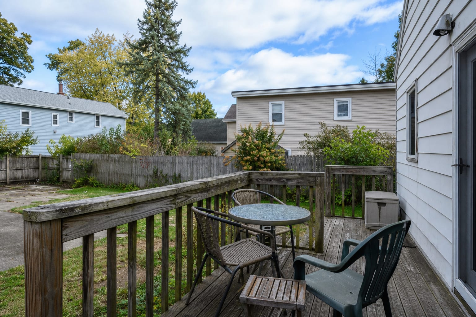 raised deck with thick dated wood railing, faded balusters, blocked yard views, and scattered furniture near the stairs.