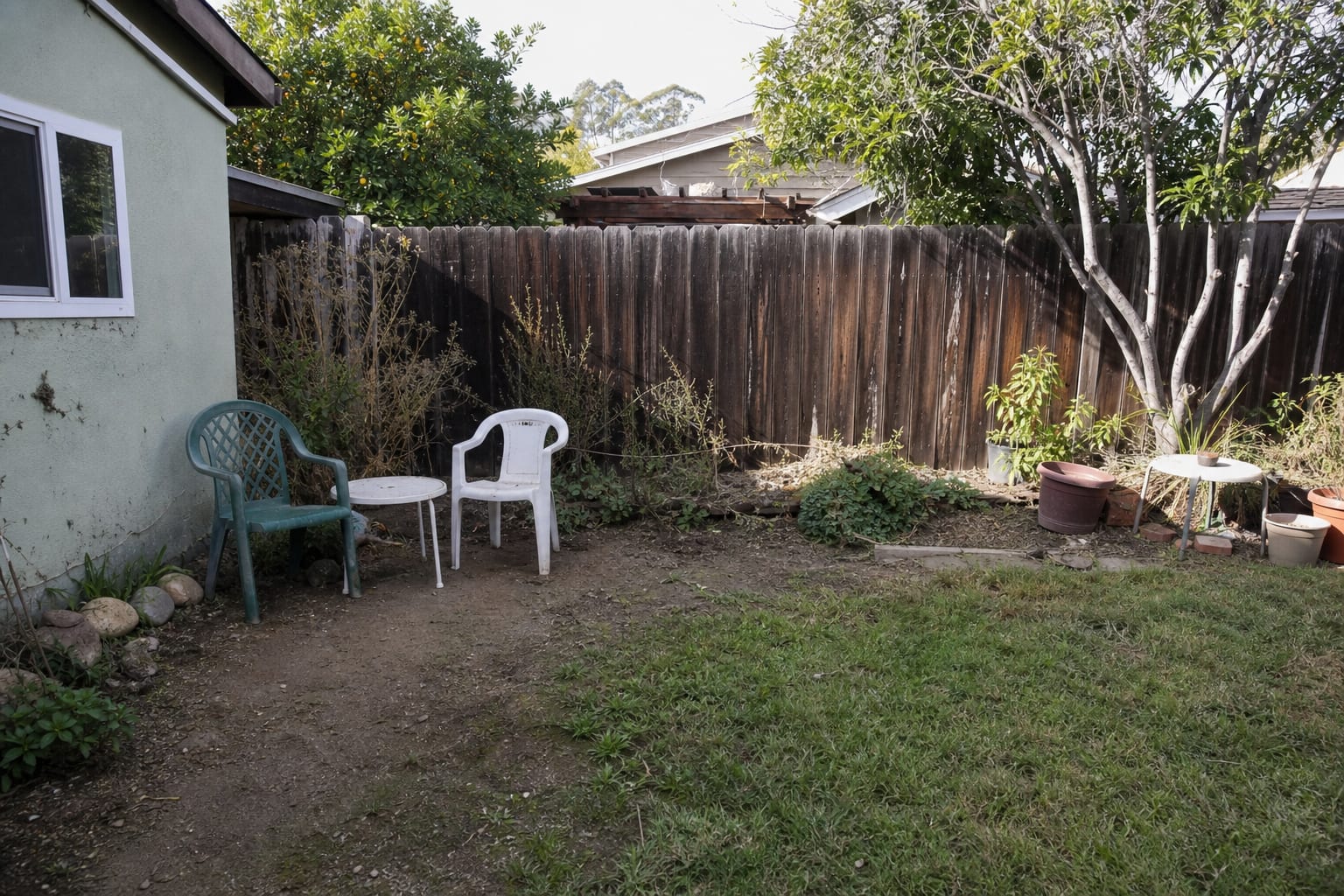 patchy dirt patio area with scattered chairs, no defined border, exposed soil, and an unfinished garden edge beside the house.