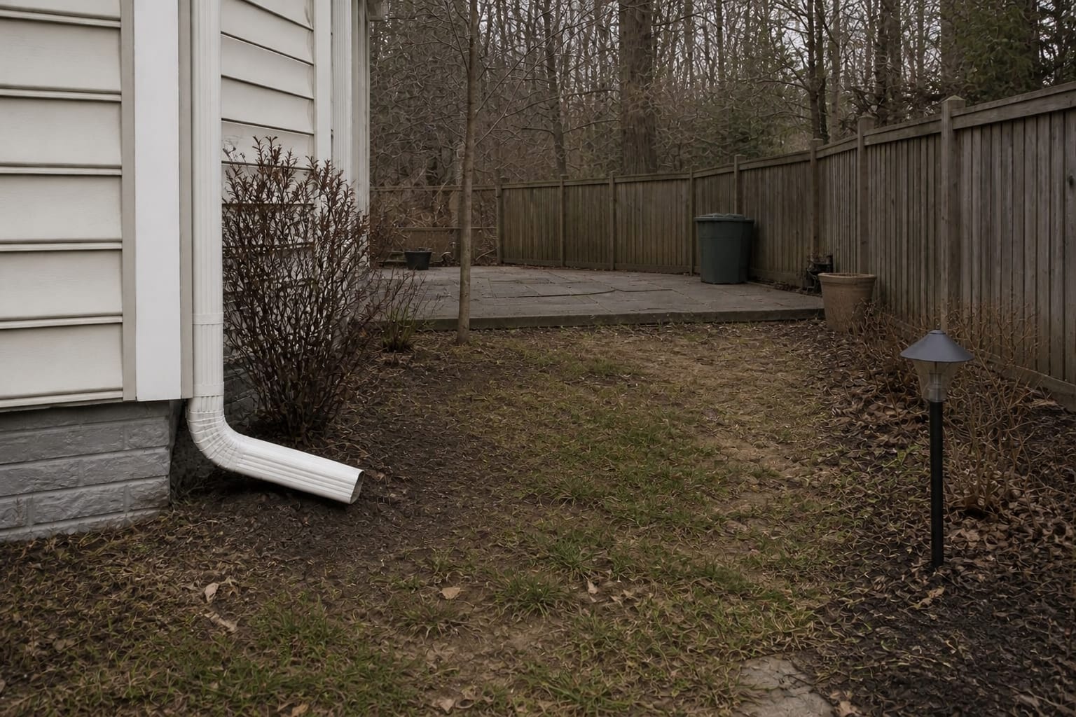 Plain downspout emptying into eroded soil beside a garden bed and foundation wall