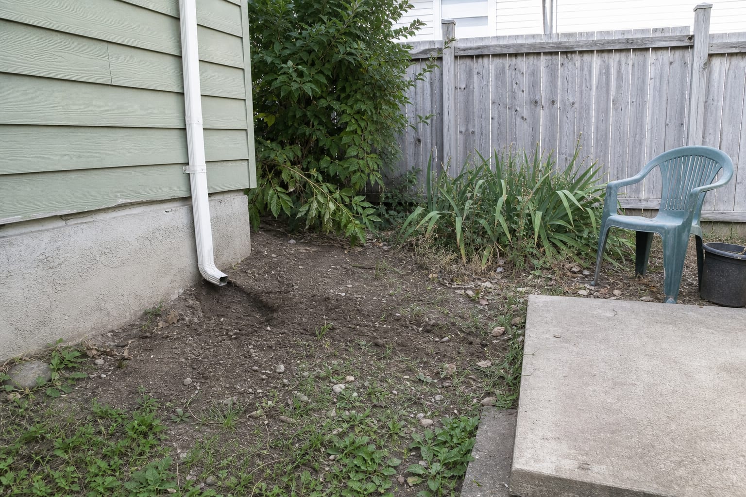 Plain downspout emptying into eroded soil beside a garden bed and foundation wall