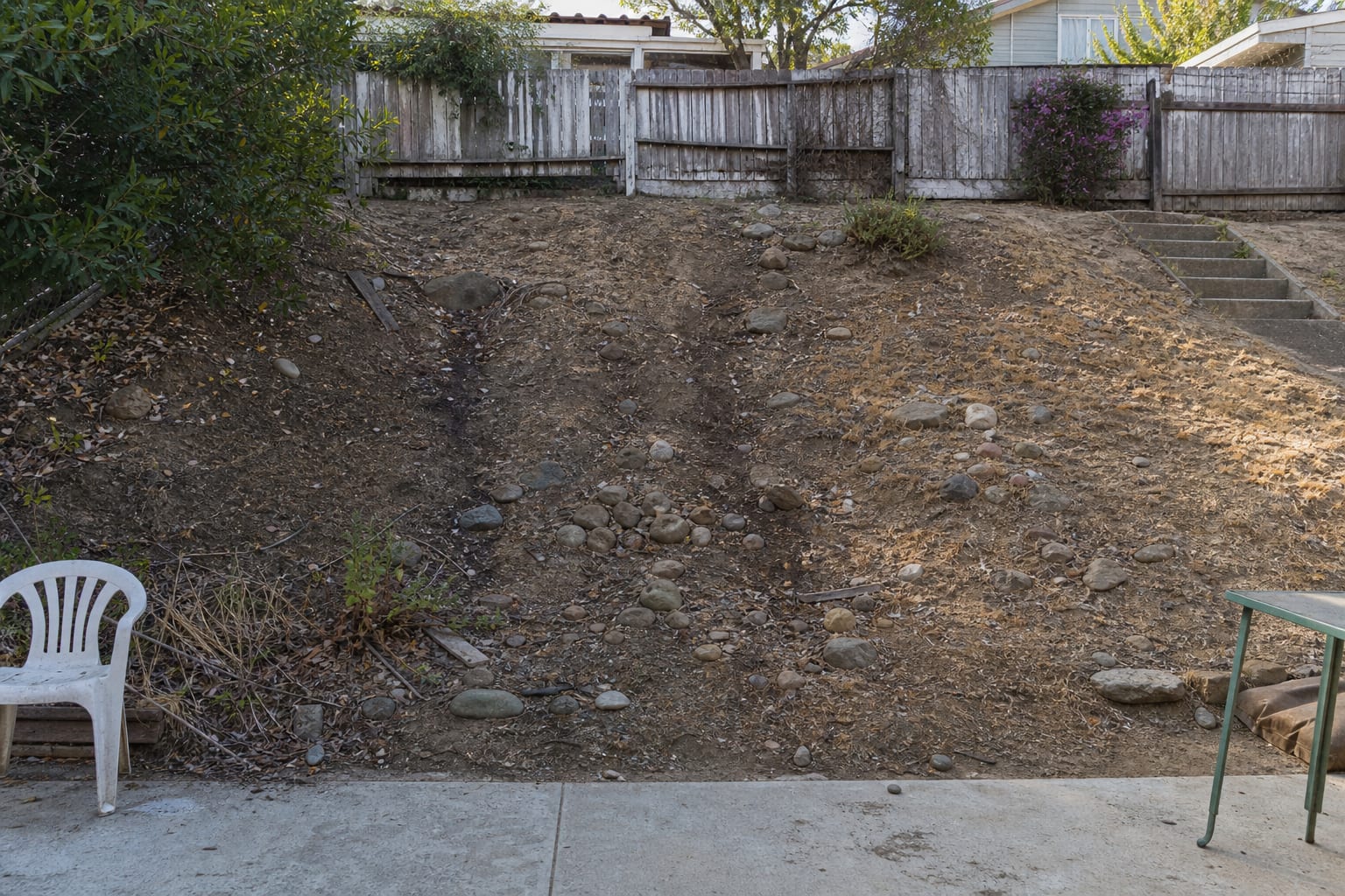 sloped backyard planting bed with exposed soil, runoff stains, scattered small rocks, and no defined drainage path