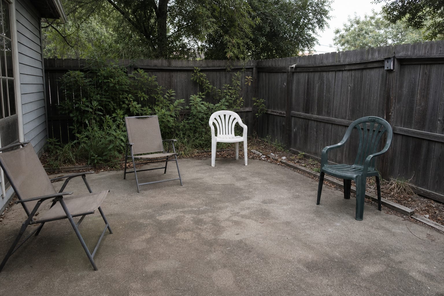 plain backyard patio corner with scattered folding chairs, a bare concrete slab, no lighting, and no defined fire pit zone
