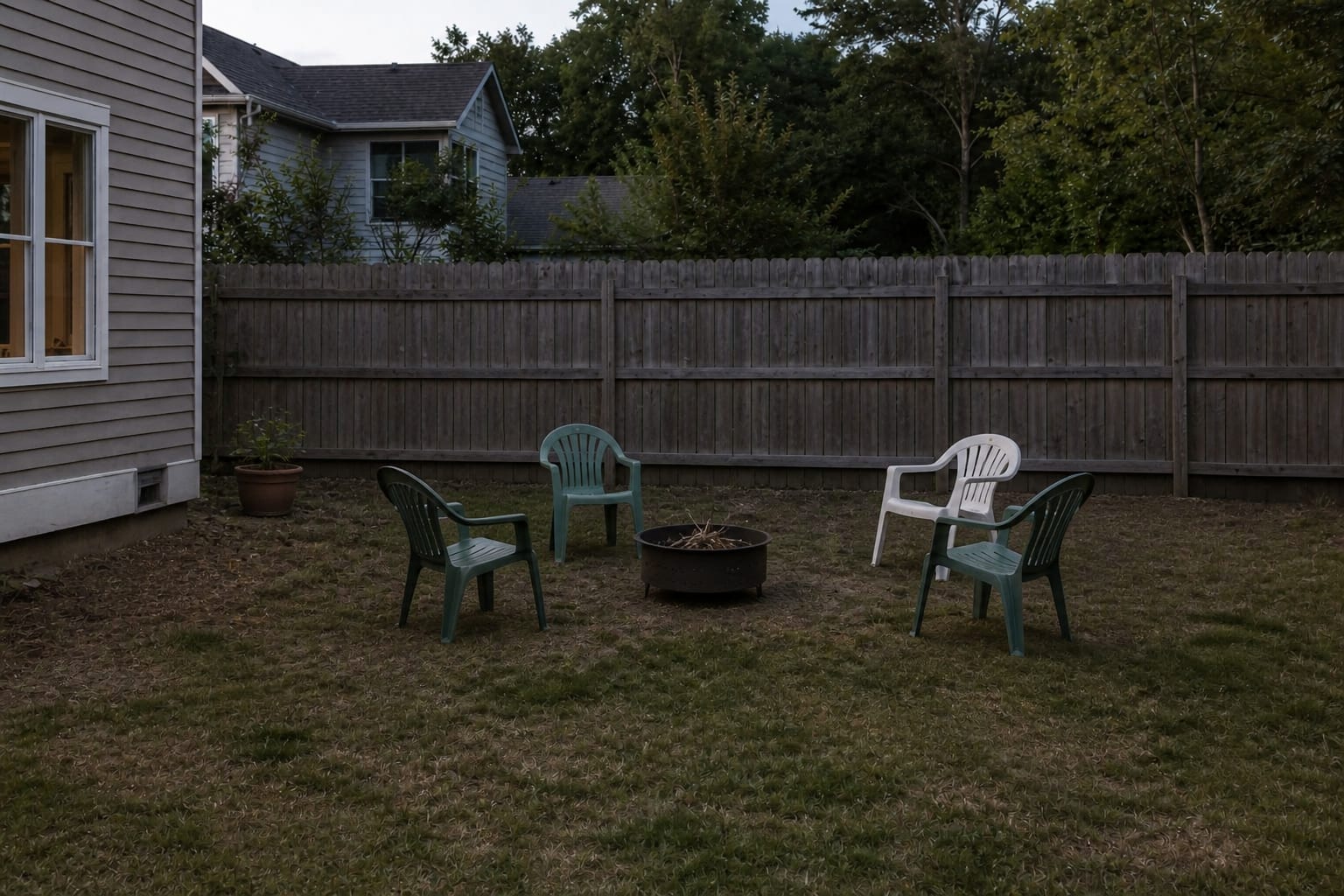 lonely metal fire pit on patchy lawn with scattered folding chairs, no defined path, and an exposed fence behind it