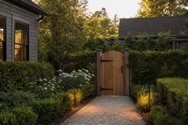 cedar garden gate with black metal hardware, gravel path, clipped hedging, and warm low lights marking the entrance