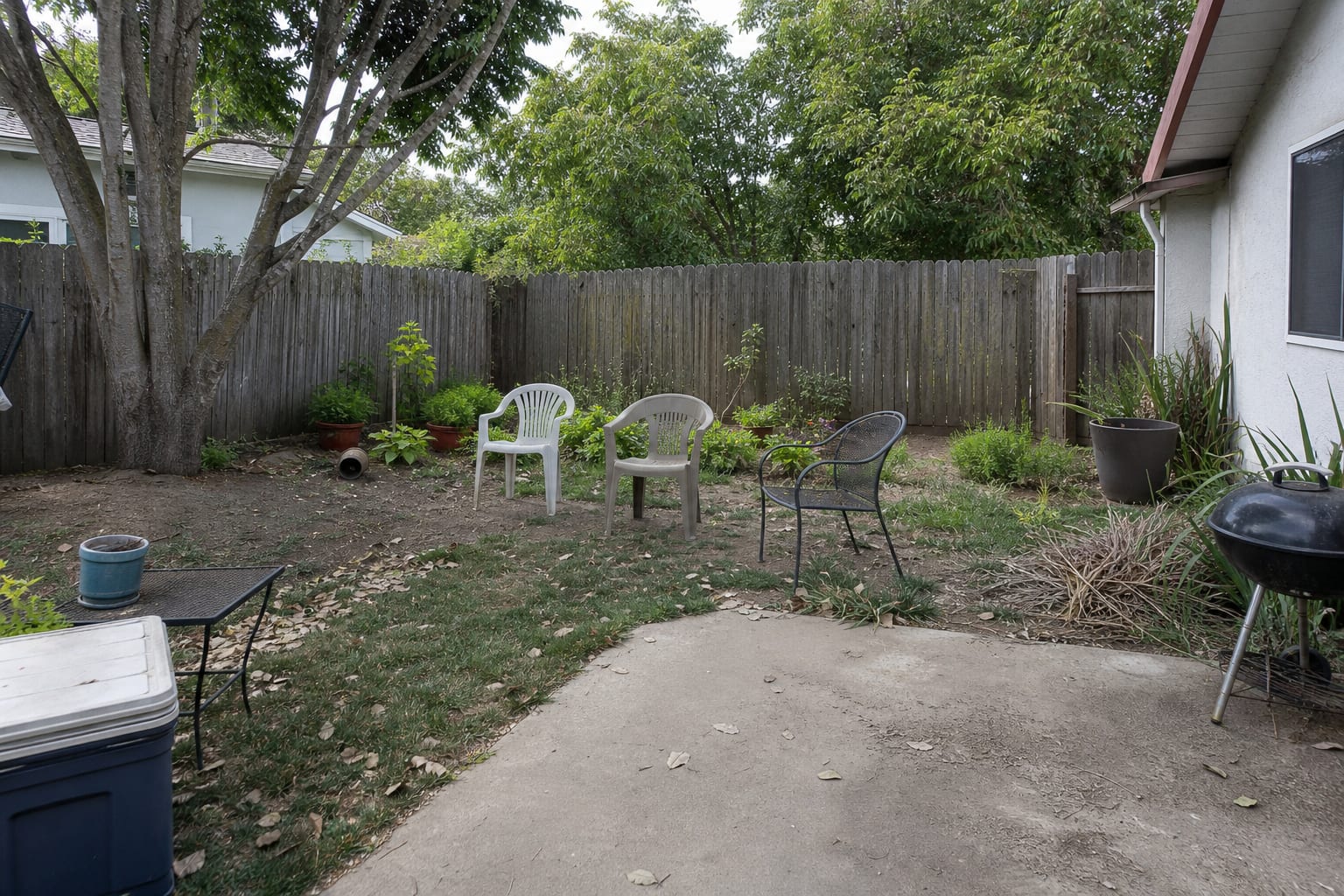 patchy lawn corner with bare soil, scattered chairs, no defined patio edge, and a blank fence behind the seating area.