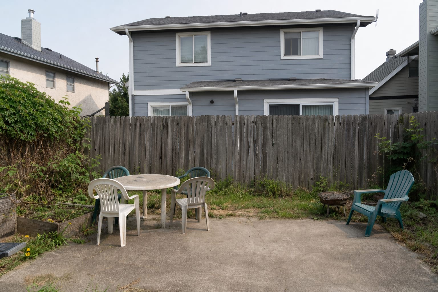 backyard with dated vertical fence panels, patchy grass, exposed neighboring view, and mismatched patio furniture near the boundary.
