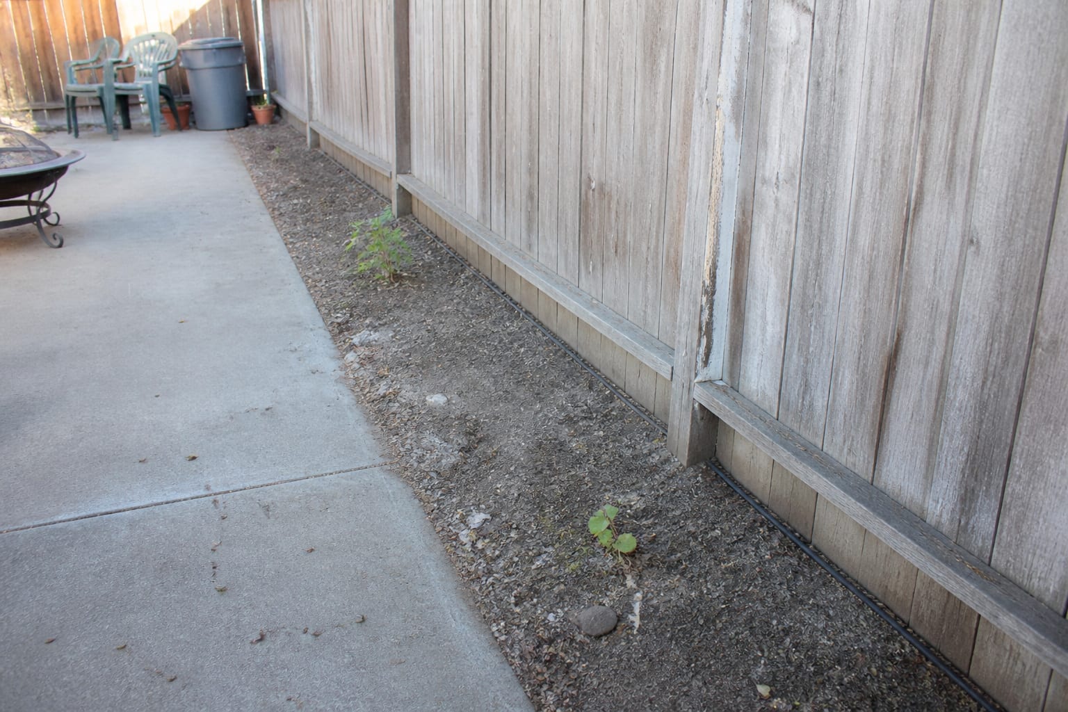 Empty narrow bed of bare soil along a fence with a few struggling plants before mixed border planting