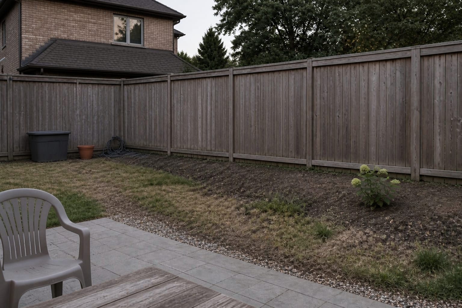 Under designed garden fence line with patchy grass, exposed soil, one small hydrangea, scattered pots, and no clear border depth or planting rhythm.