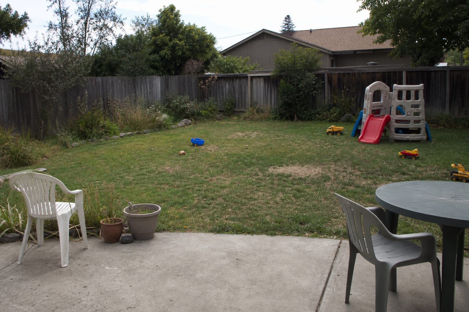 Patchy backyard lawn with loose toys, no shade, and a playset pushed awkwardly against the fence.