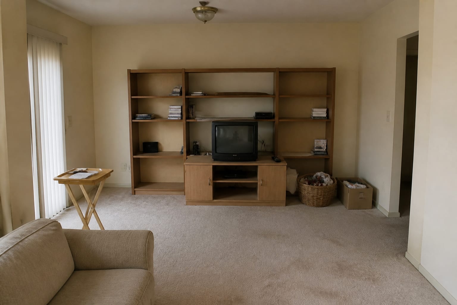 Living-room wall covered in a mismatched étagère, low maple TV stand, basket of toys overflowing, cardboard bins, and a folding tray table with mail.