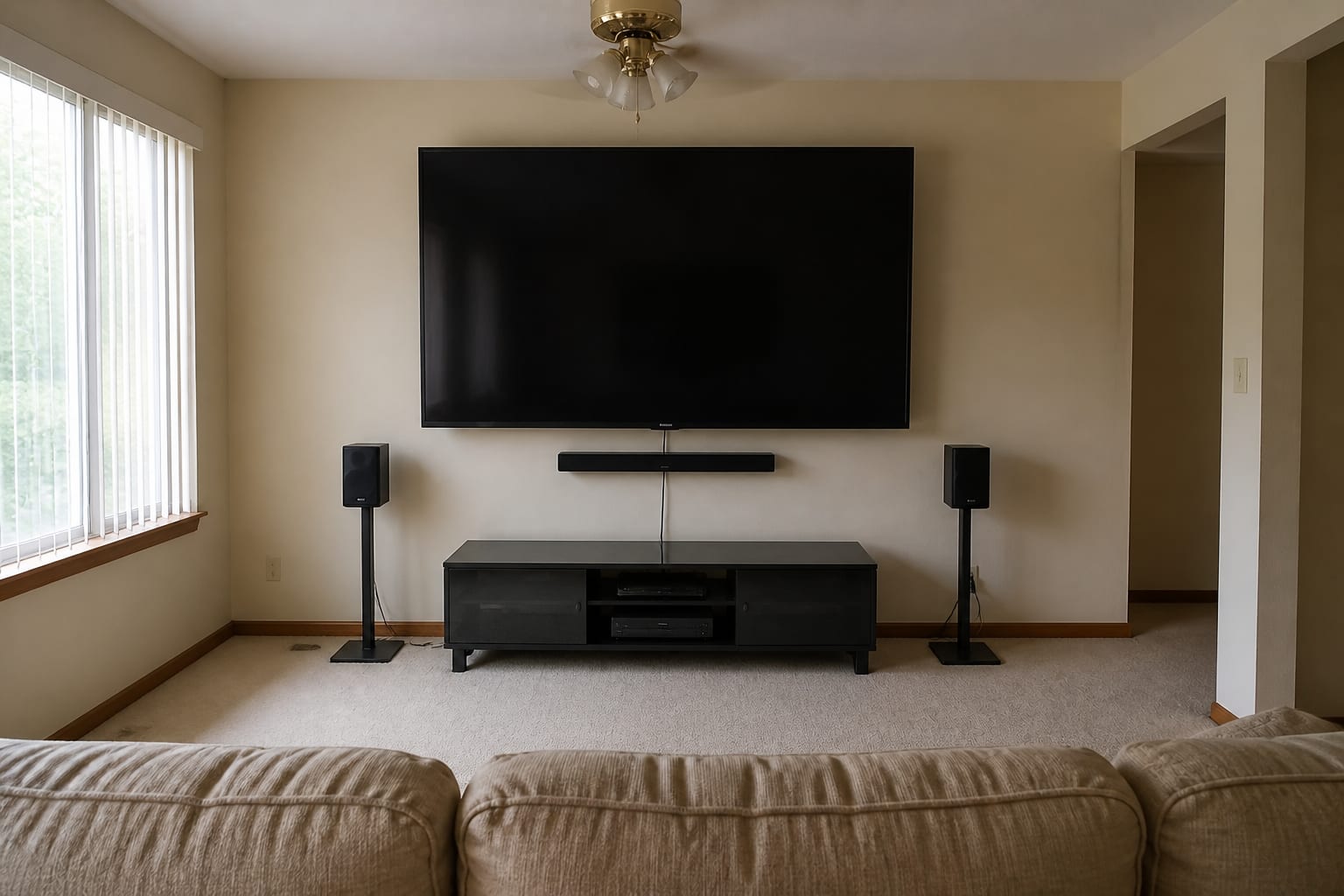 Living-room wall dominated by an oversized 85-inch TV mounted too high on a glaring white wall, a black-glass entertainment center, cords, a soundbar, and matching black speaker stands.