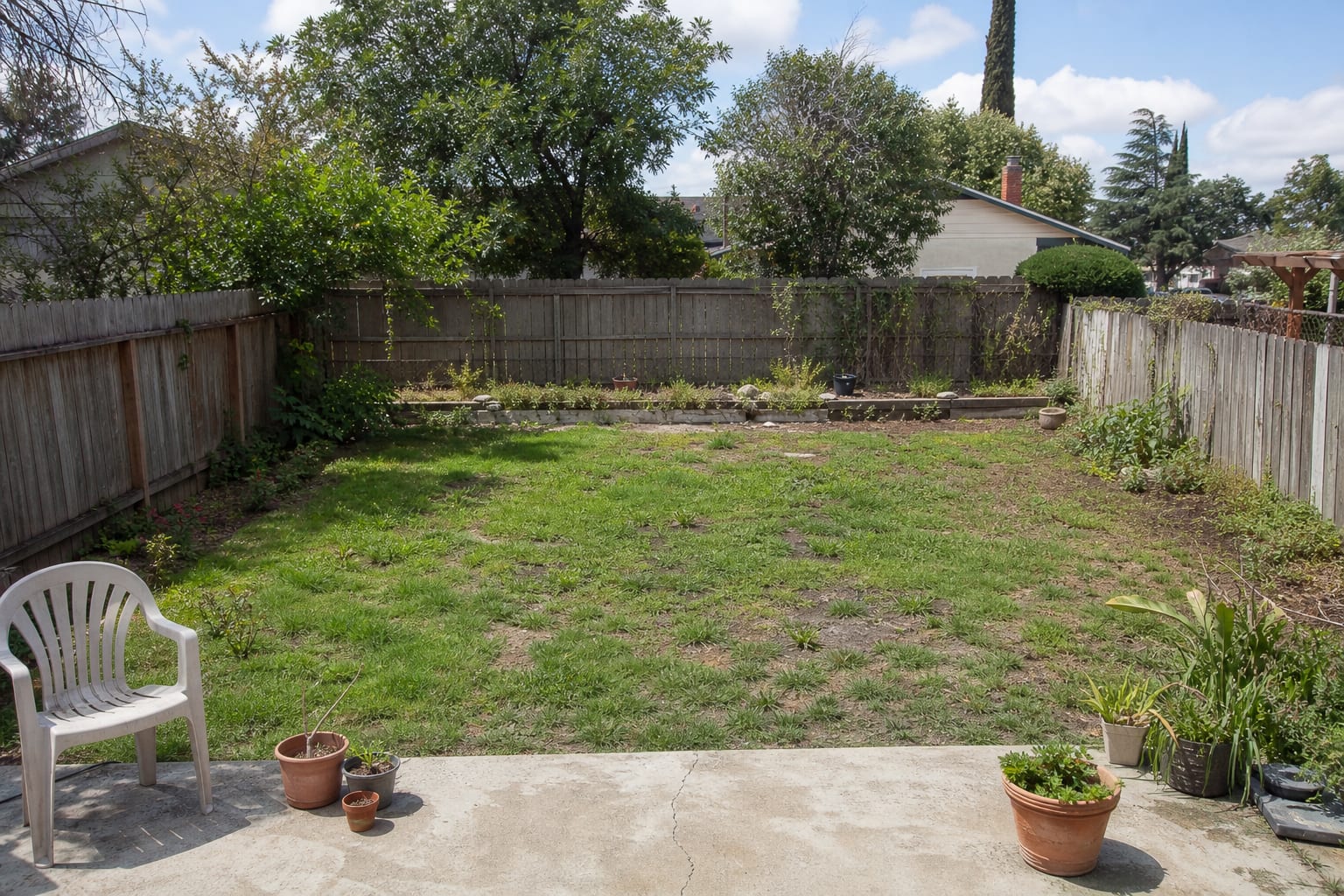 Under-designed backyard with patchy lawn, narrow planting strips, scattered pots, exposed soil, and no clear path or seating area.