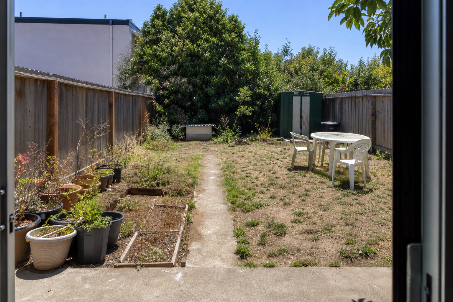 Patchy lawn garden with scattered plastic pots, exposed fencing, no shade, and an unused corner beside a narrow concrete path.