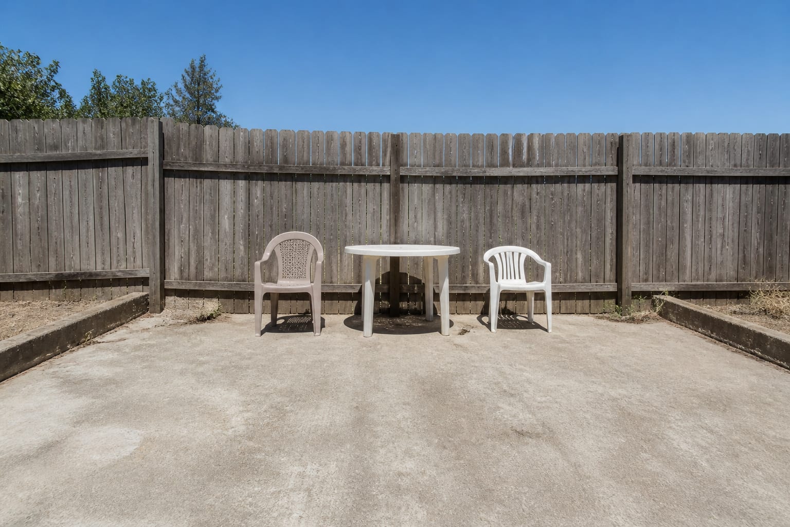 Plain concrete backyard patio with a basic plastic table, no shade, and bare fencing under flat sunlight