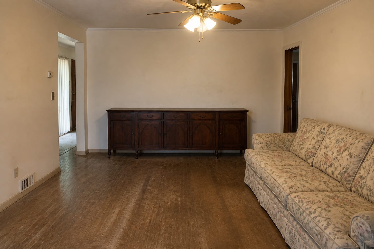 Living room with a cluttered antique sideboard, a dated floral sofa, scuffed beige walls, and harsh overhead light.