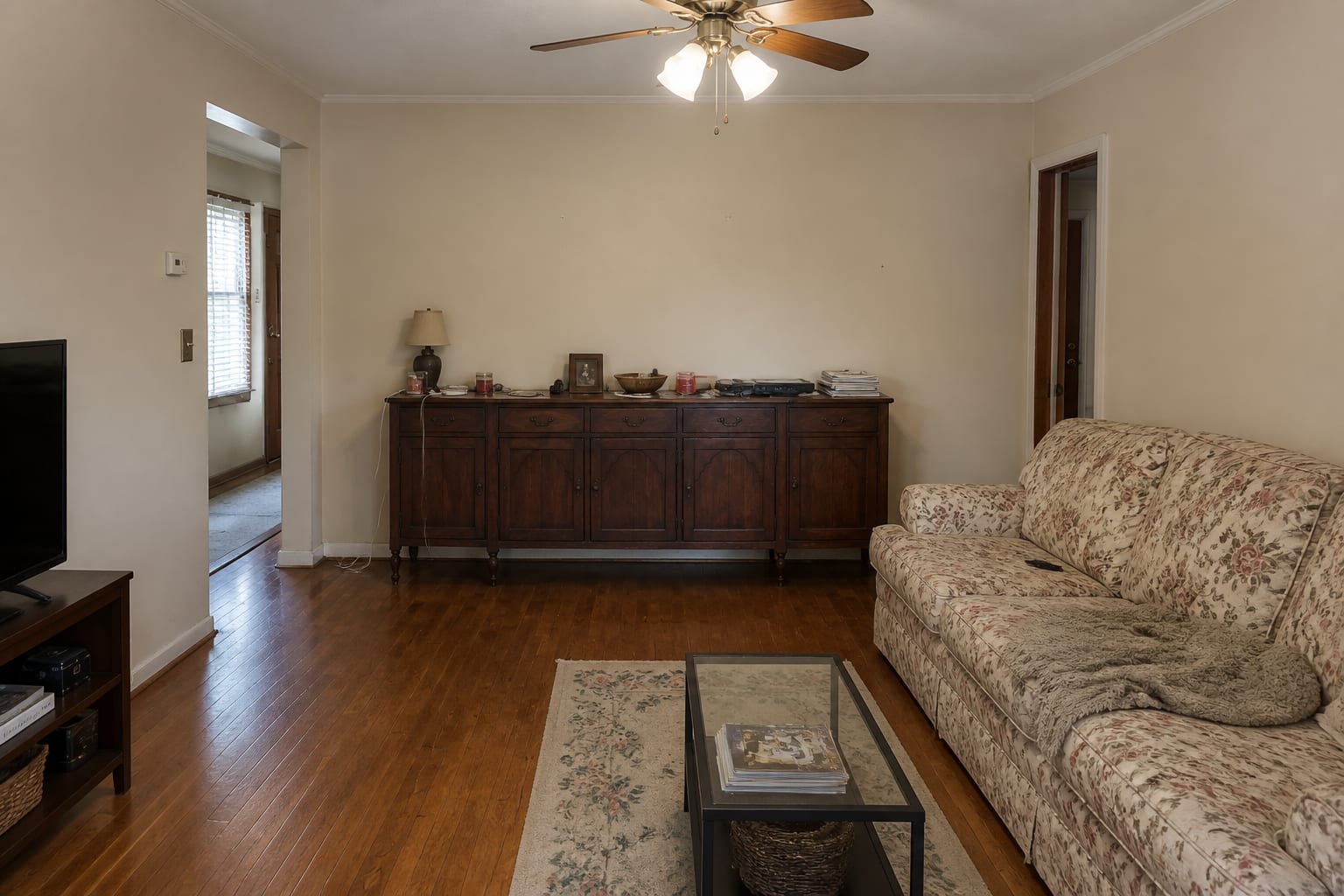 Living room with a cluttered antique sideboard, a dated floral sofa, scuffed beige walls, and harsh overhead light.
