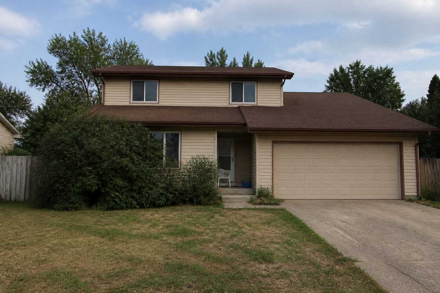 A plain suburban two-story house with faded beige siding, dated brown trim, white aluminum windows, and a bare entry before any redesign.