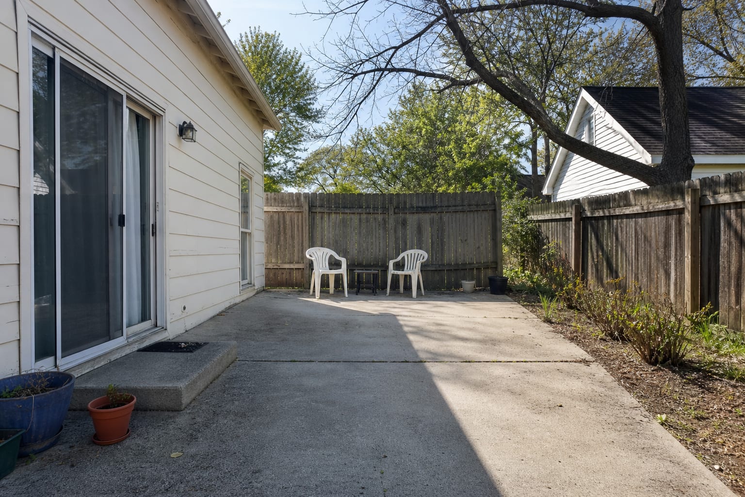 under-designed concrete patio with small scattered chairs, exposed siding, no shade, no rug, and a few undersized pots near the door.