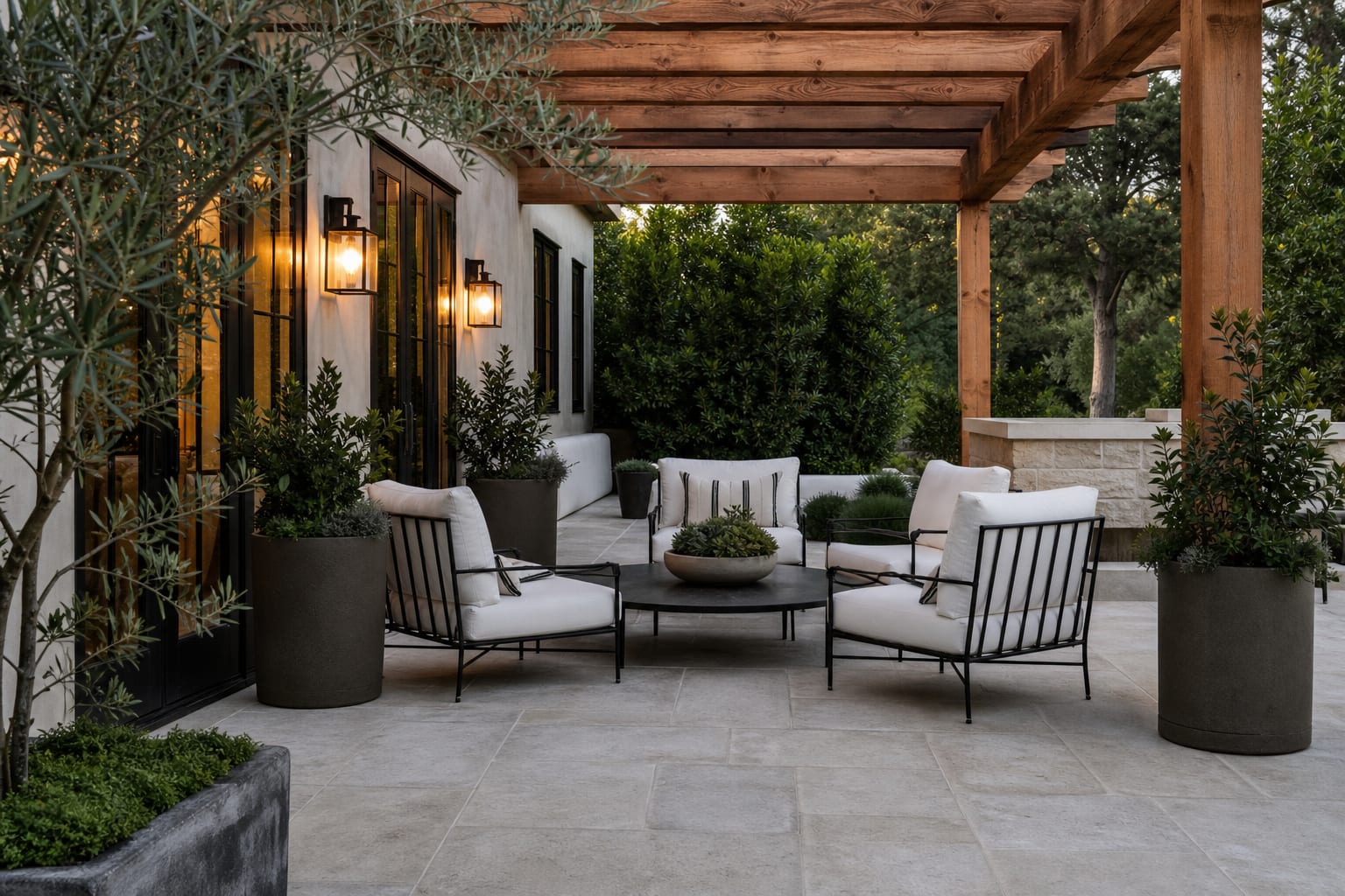 modern farmhouse patio with black metal chairs, pale stone pavers, cedar pergola, olive planters, and warm sconces