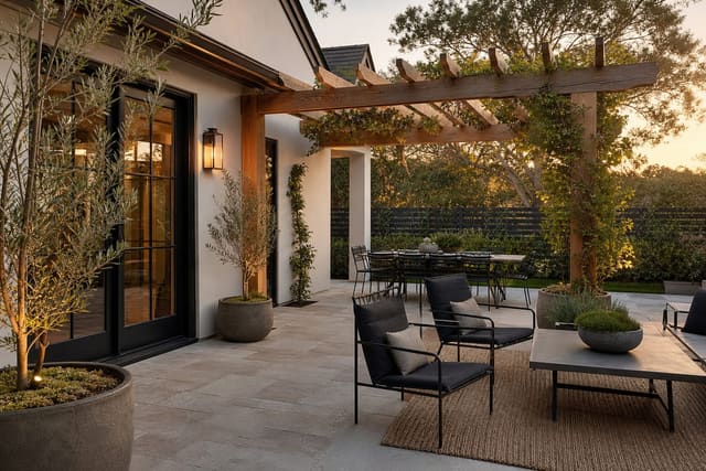 modern farmhouse patio with black metal chairs, pale stone pavers, cedar pergola, olive planters, and warm sconces