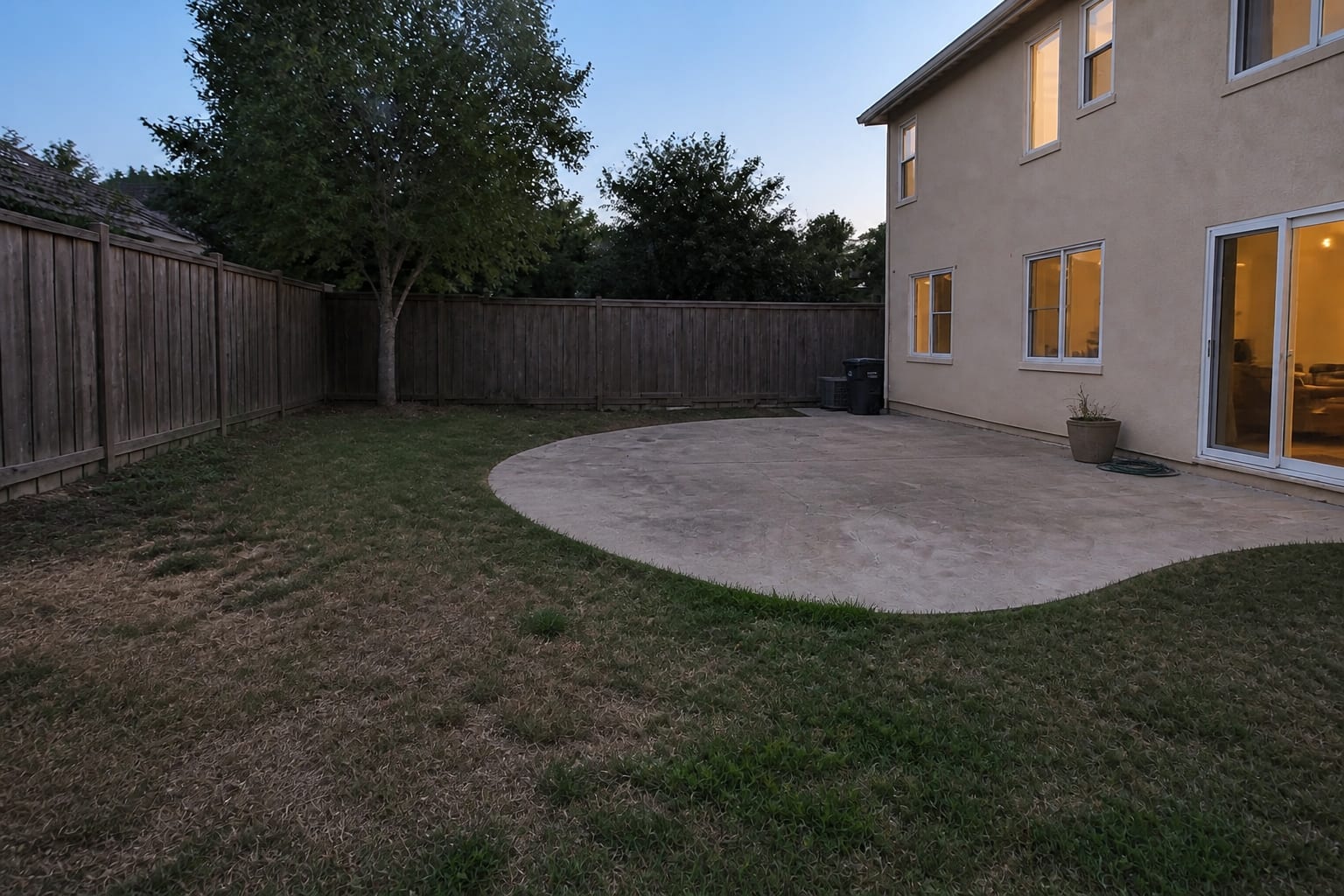 A plain suburban backyard with a flat green lawn and an empty gray concrete patio before redesign